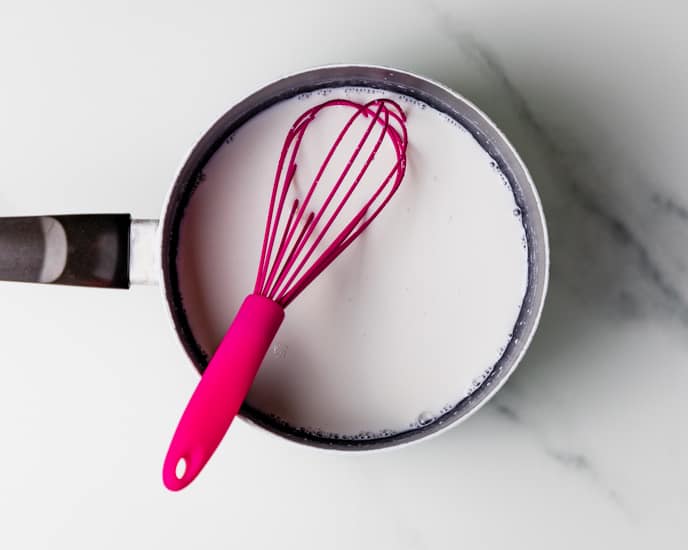 Milk, water and flour in a saucepan with a pink whisk. 